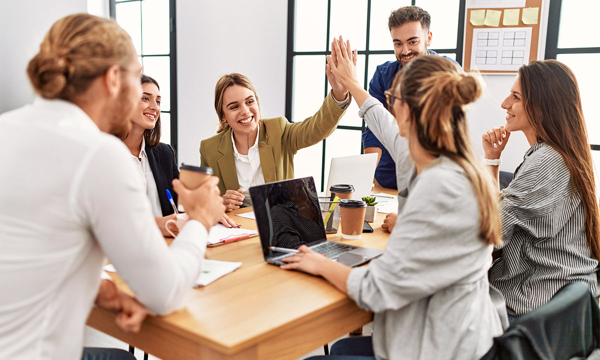 Mehrere Personen sitzen an einem Tisch in einer freundlichen Arbeitsumgebung. Zwei geben sich ein High-Five.